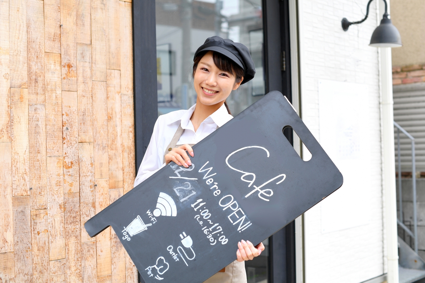 カフェの店頭で黒い色の店頭POPを両手で持って微笑んでいる白いシャツの女性店員