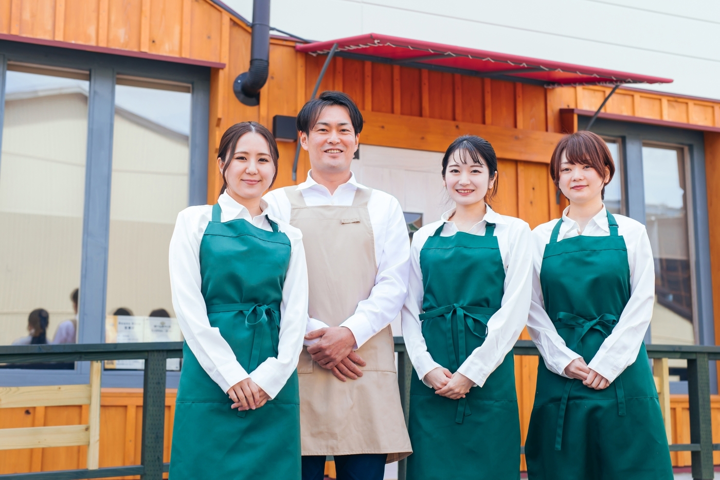 お店の前に並ぶ白いシャツを着て緑のエプロンを着用した3人の若い女性スタッフと、白いシャツを着てベージュのエプロンを身に着けた1人の男性スタッフ