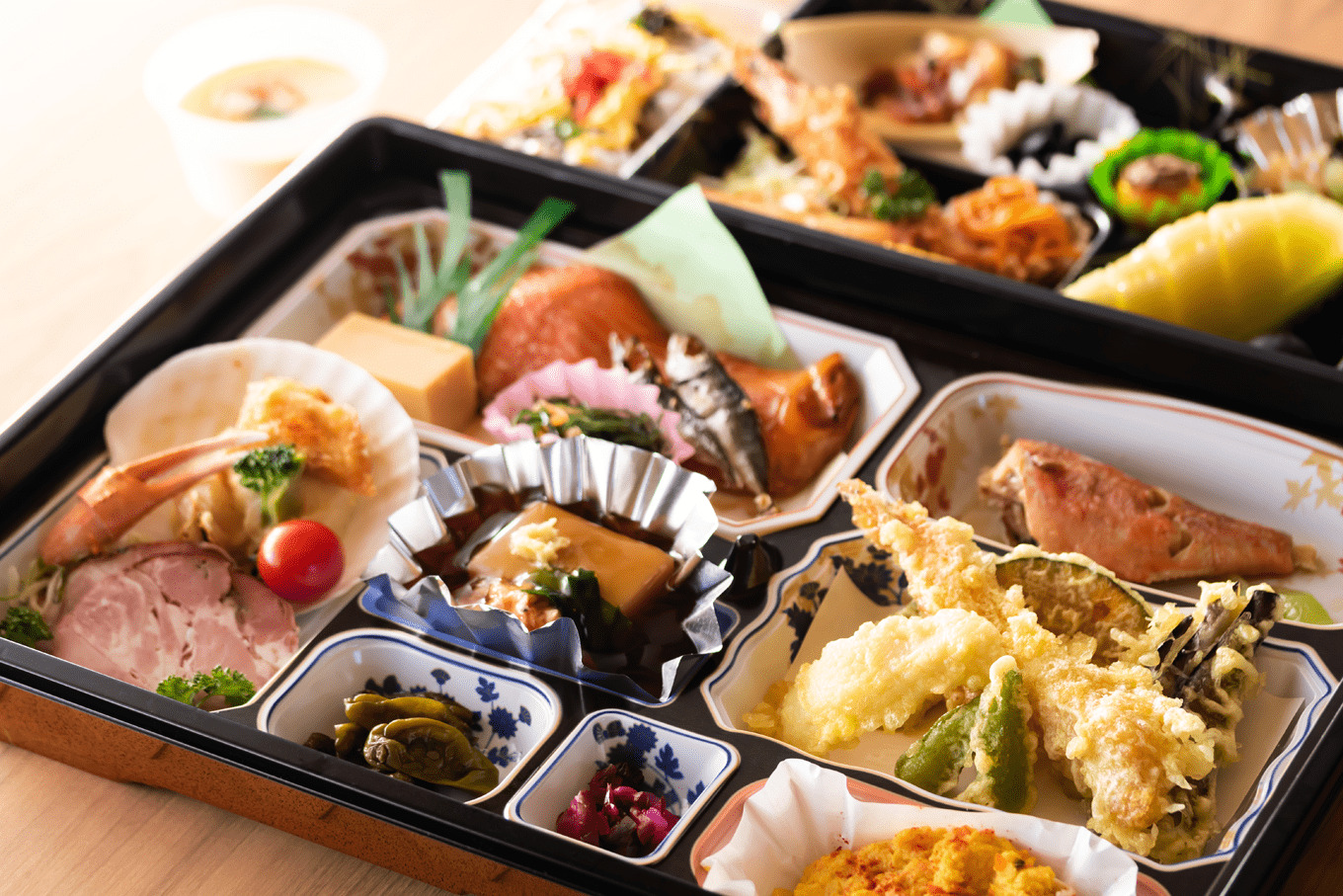 お弁当の容器の中に小皿がたくさんあり、焼き魚や天ぷら、漬物など多くの料理が盛りつけされている。
